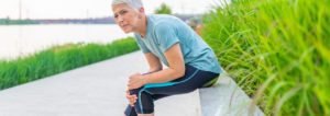An older female jogger sits on bench clutching her knee in pain.