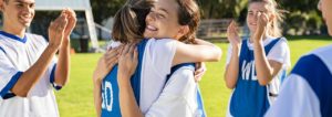 Two girls in netball uniform hug each other as their peers look on clapping.