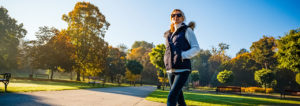 A lady wearing warm activewear walks through a sunny and leafy green park, with her hands in her pockets.