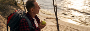 Man with a backpack, holding a green apple and staring into the distance.