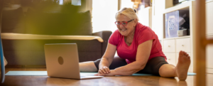 A woman in activewear smiles at laptop while stretching on the floor