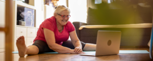 A woman in activewear smiles at laptop while stretching on the floor